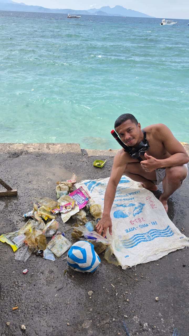 Young people collecting plastic waste and debris on a sandy beach with the ocean and distant trees visible. The mood is determined and community-focused.